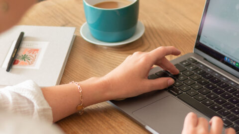 Person working on laptop at desk with coffee cup and notebook.