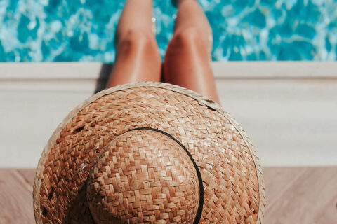 Person sitting poolside in straw hat with their feet in the water.