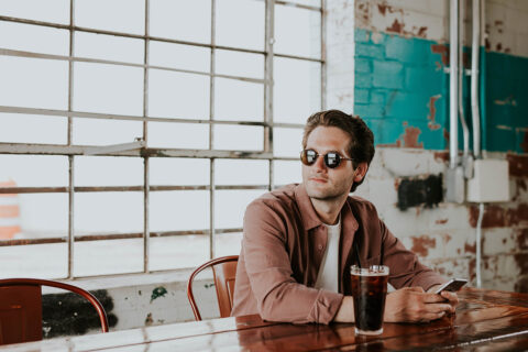 Person in sunglasses sitting at coffeeshop table with smart phone and iced coffee.