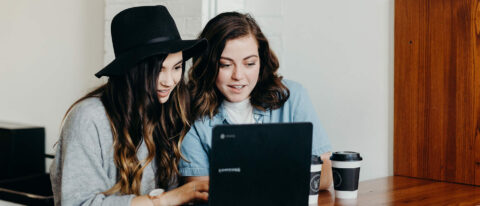 Two friends working together on computer at coffee shop desk.