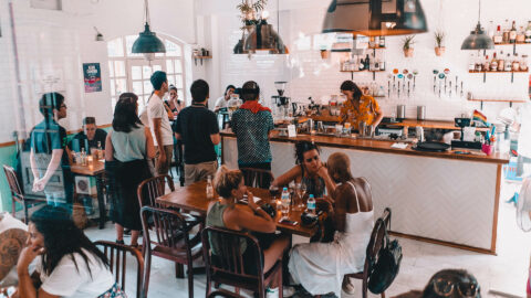 Busy coffee shop and bar with white tiled walls, shelves of liquor, and beer taps.